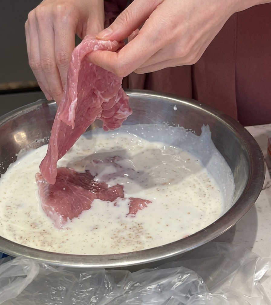 Des mains plongeant une fine tranche de porc rose cru dans un bol en inox rempli dune marinade blanche au lait et à lail.
