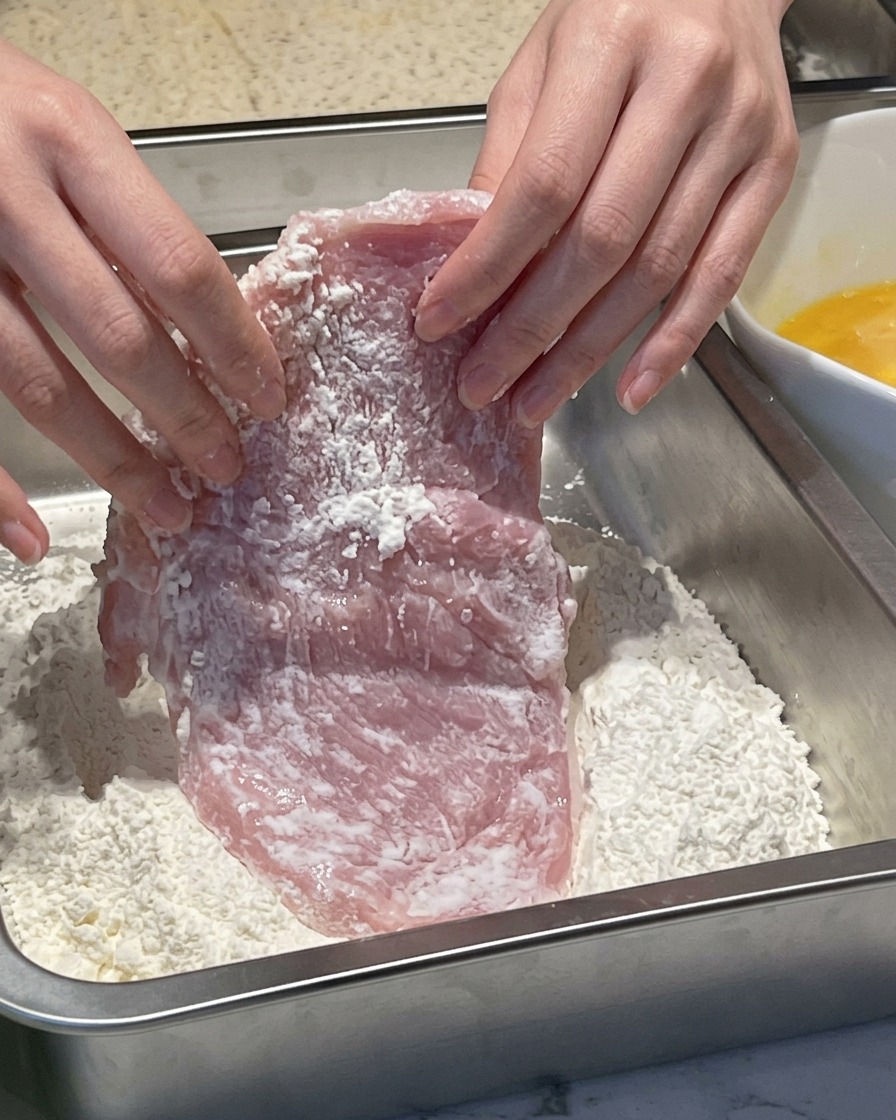 Des mains pressant un morceau de porc humide dans un récipient métallique rempli de farine blanche.