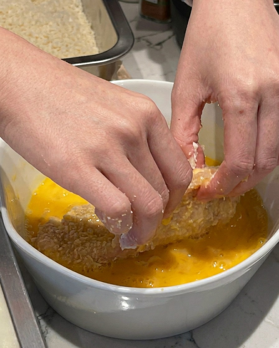 Une main trempant une escalope de porc farinée dans un bol en céramique blanche contenant des œufs battus jaunes.