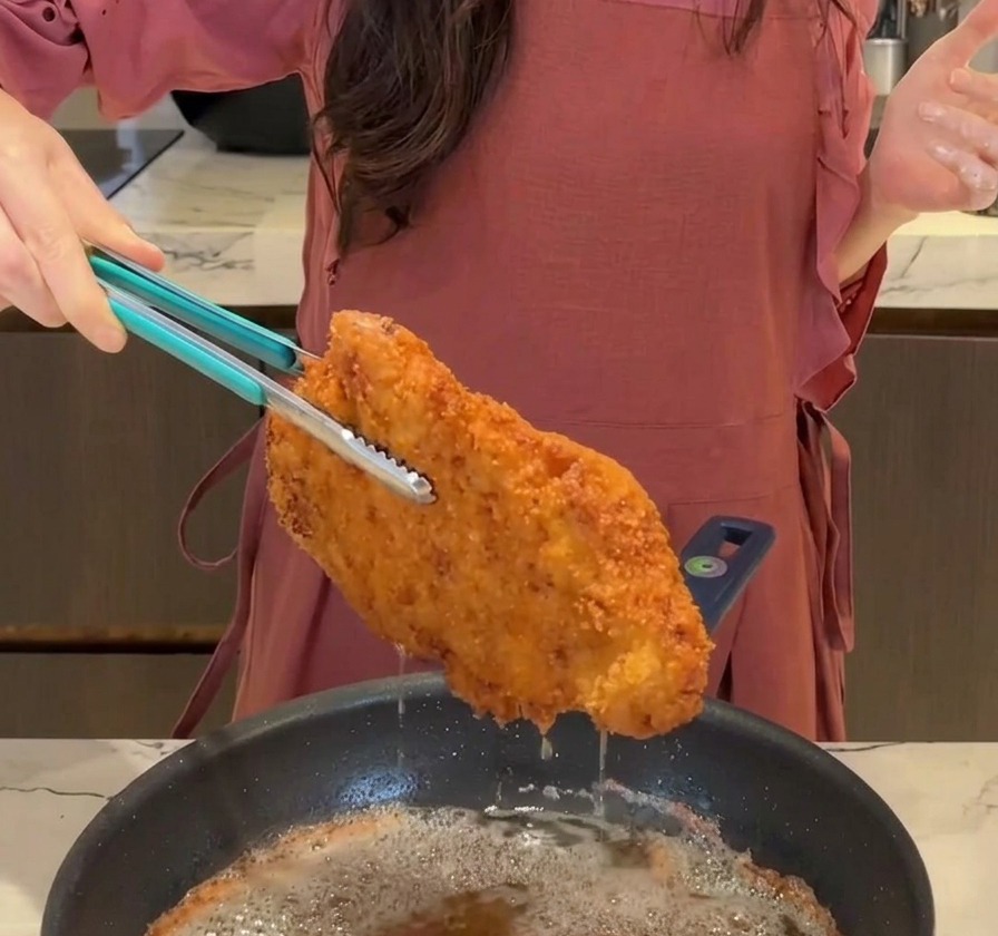 Une grande escalope de porc frite, dorée et croustillante, soulevée de la poêle avec une pince bleue, laissant lhuile ségoutter.