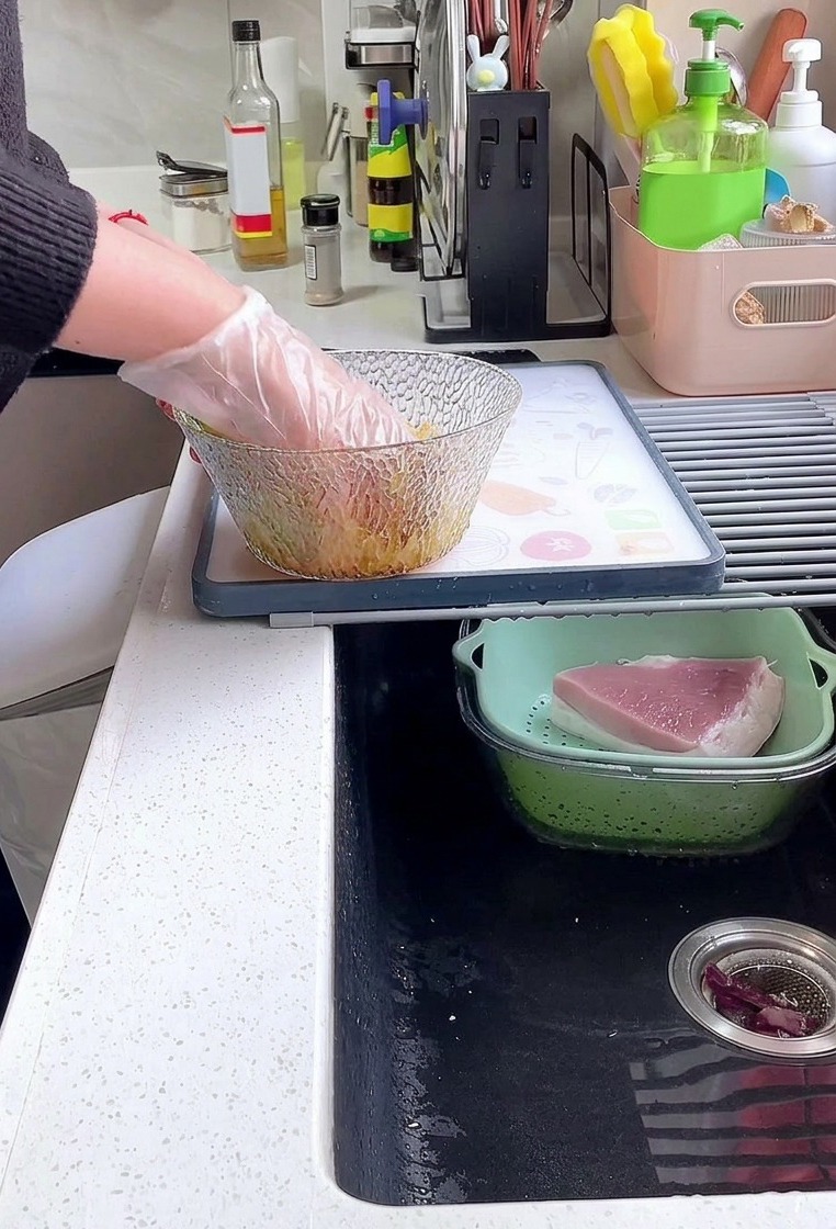 Une personne avec un gant en plastique mélangeant dépaisses tranches de longe de porc avec de la sauce soja, du poivre et de la fécule dans un bol en verre.