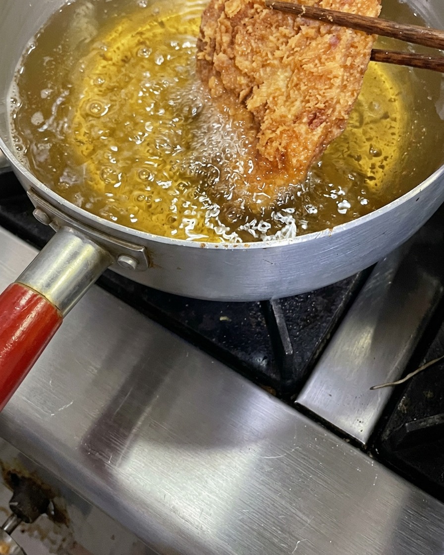 Une côte de porc tonkatsu bien dorée et croustillante soulevée avec des baguettes en bois au-dessus dune casserole dhuile de friture bouillonnante.