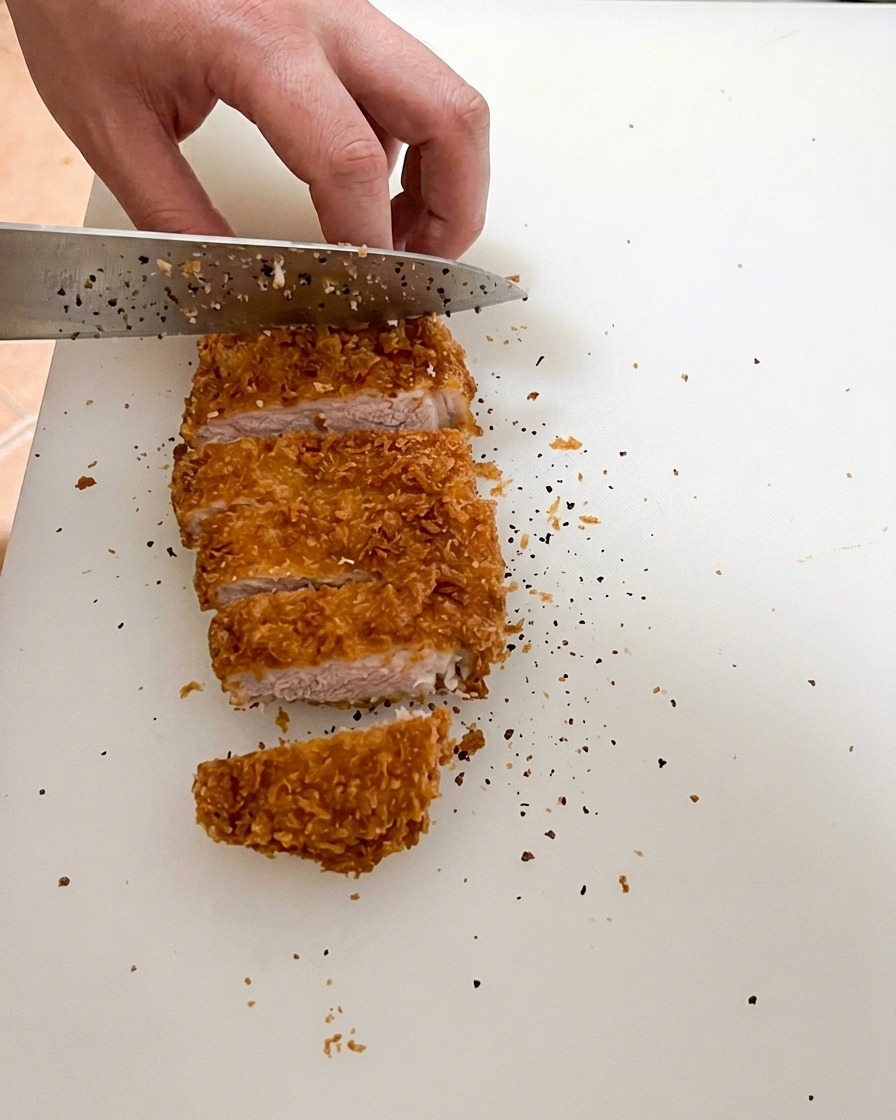 Un couteau aiguisé tranchant une côte de porc parfaitement frite sur une planche à découper blanche, révélant une viande juteuse au cœur légèrement rosé.