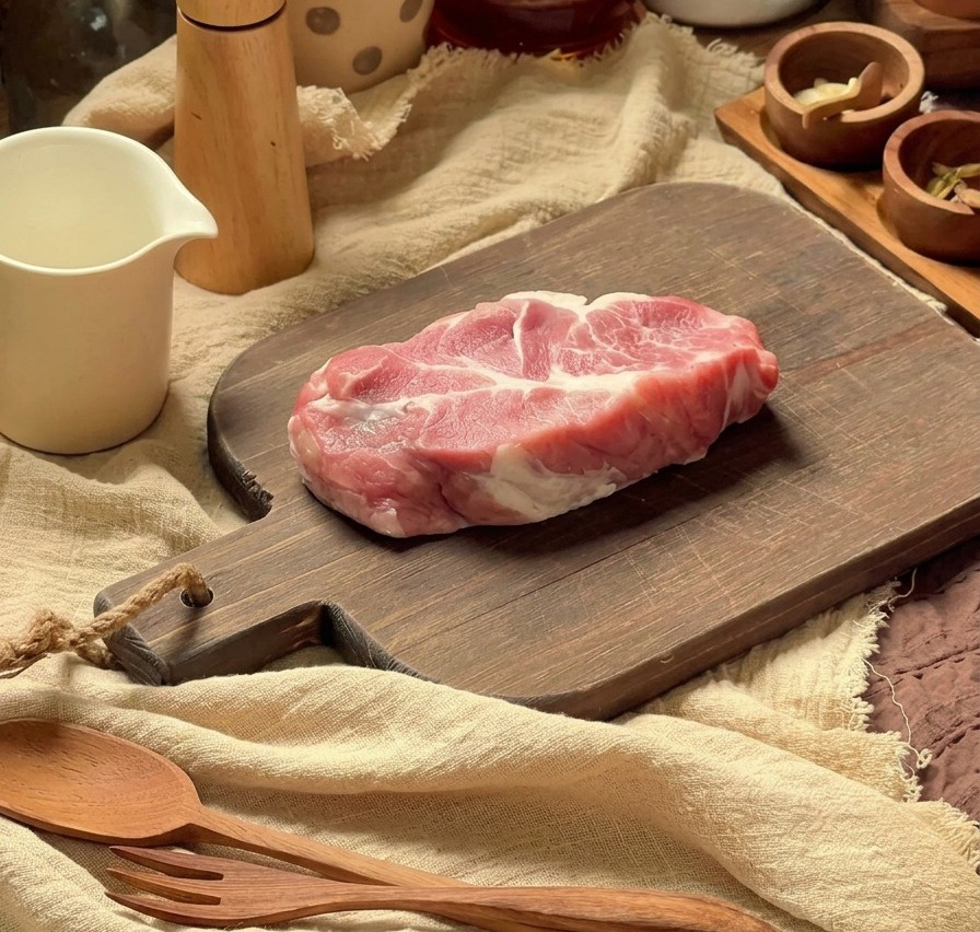Côtelette de porc crue sur une planche à découper en bois avec des ustensiles de cuisine en arrière-plan.