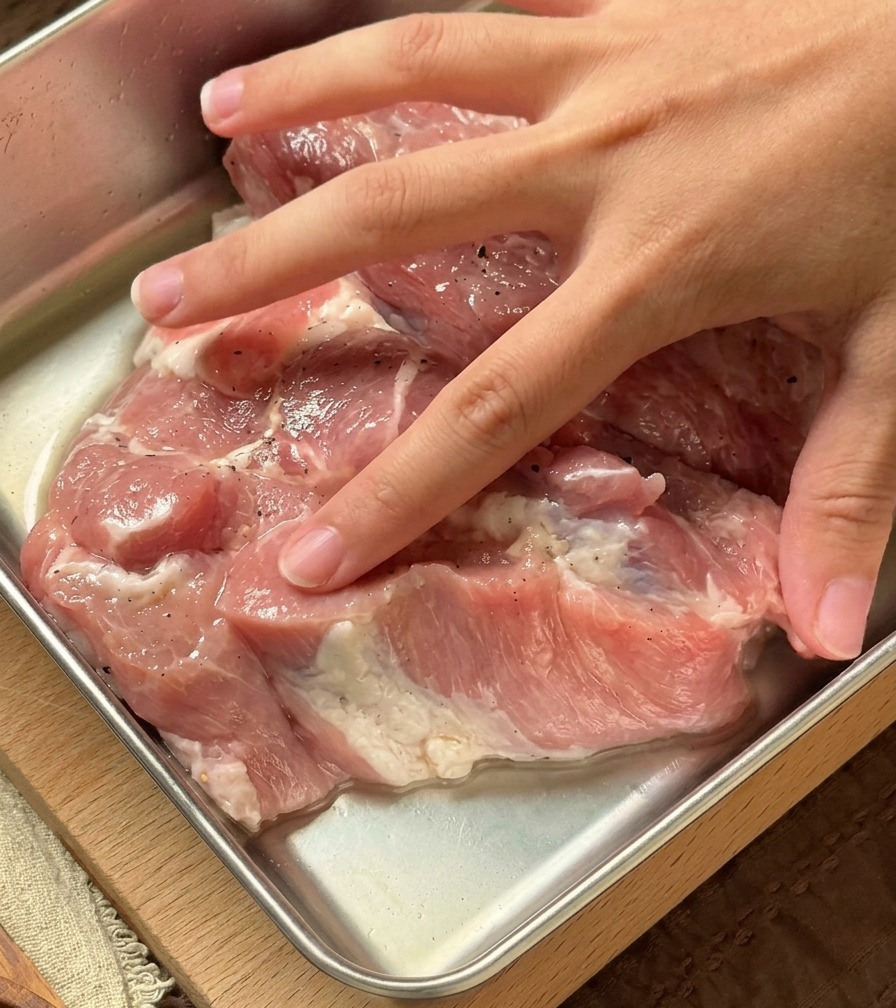Une main incorporant un liquide aromatique clair dans des côtelettes de porc crues dans un récipient métallique.