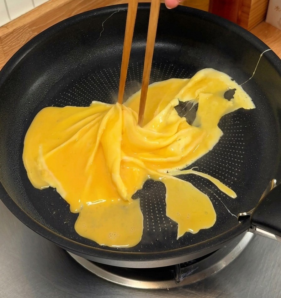 Des baguettes faisant tourner une omelette jaune à moitié cuite dans une poêle antiadhésive noire pour former une spirale.