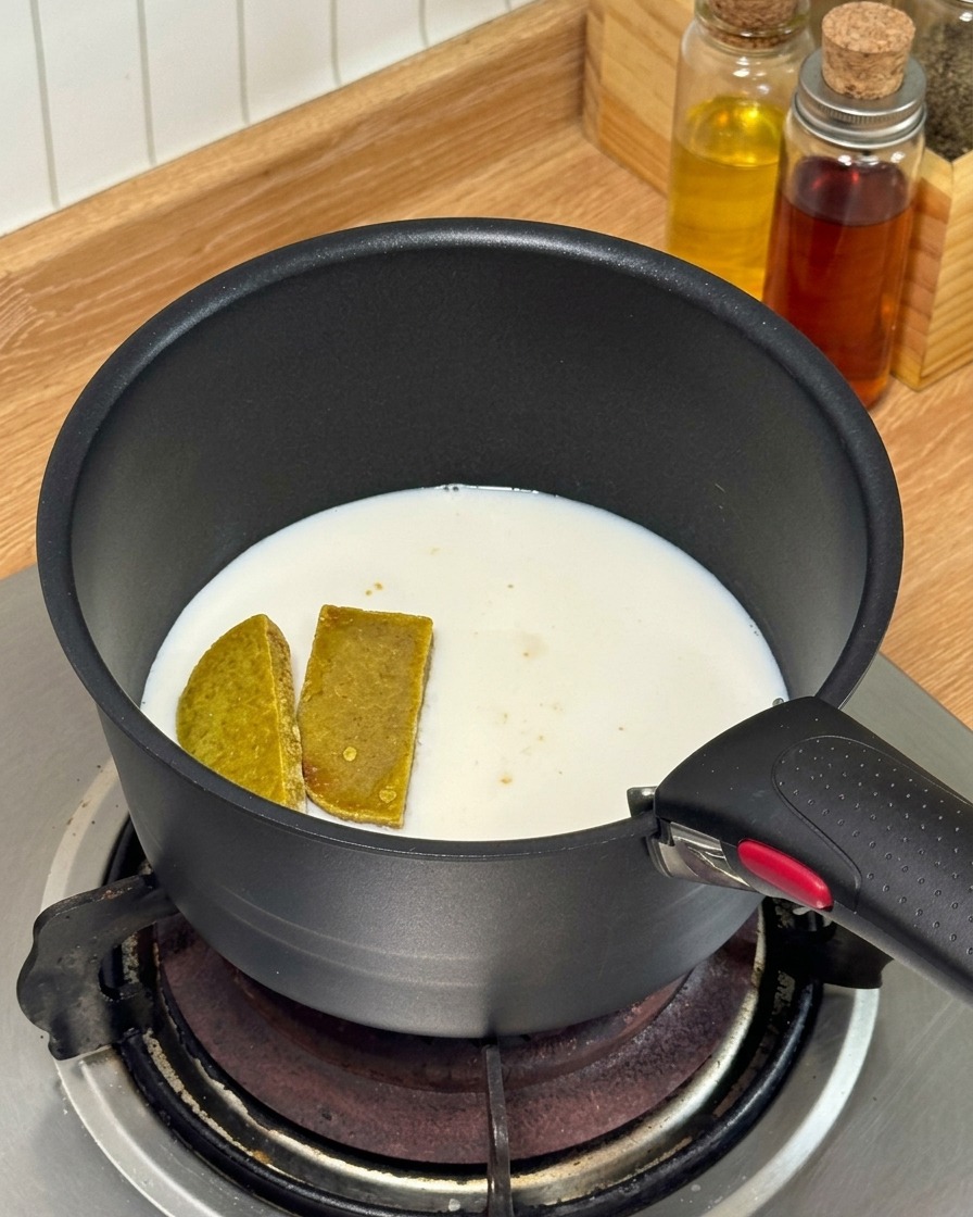 Des cubes de curry et du lait qui chauffent dans une petite casserole noire sur une cuisinière à gaz.