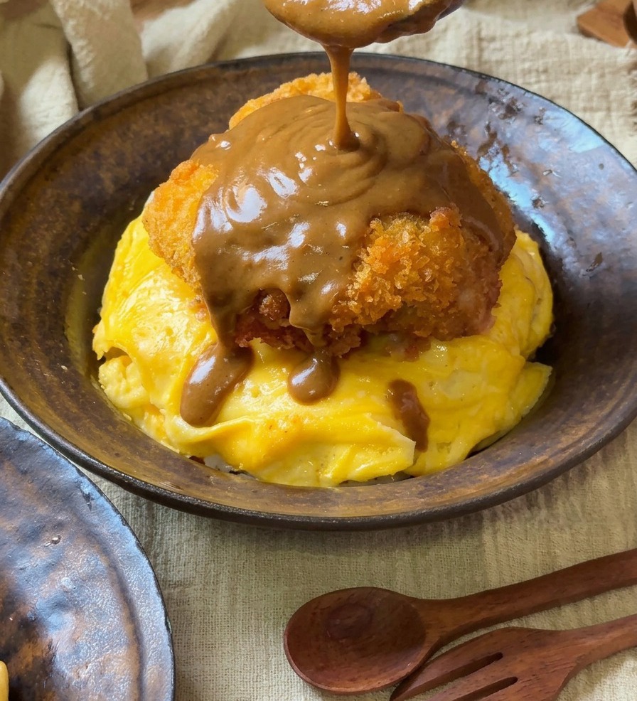 Une sauce curry brune et onctueuse versée à la cuillère en bois sur une côtelette de porc panée posée sur un nid domelette en spirale.