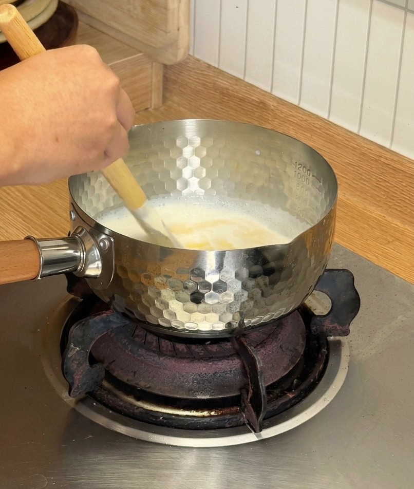 Une spatule en bois remuant un mélange blanc crémeux de fromage fondant et de lait de coco dans une petite casserole argentée.