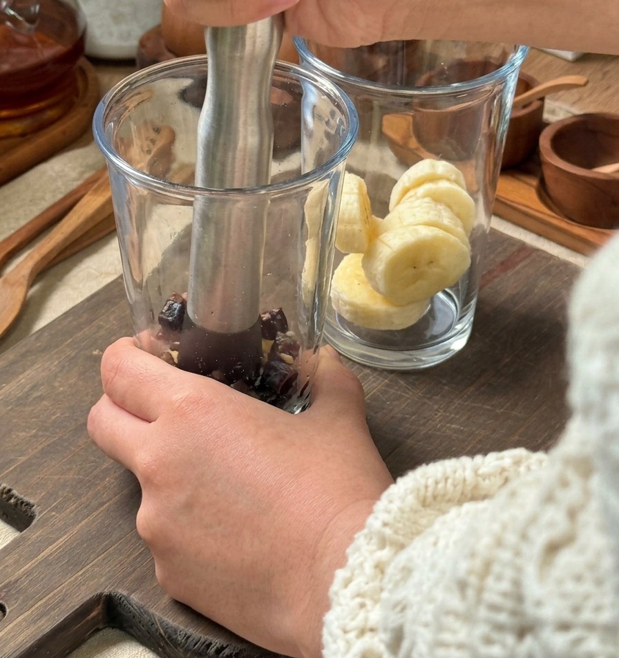 Des mains utilisant un pilon métallique pour écraser des ingrédients dans un grand verre, avec un autre verre contenant des rondelles de banane à proximité.