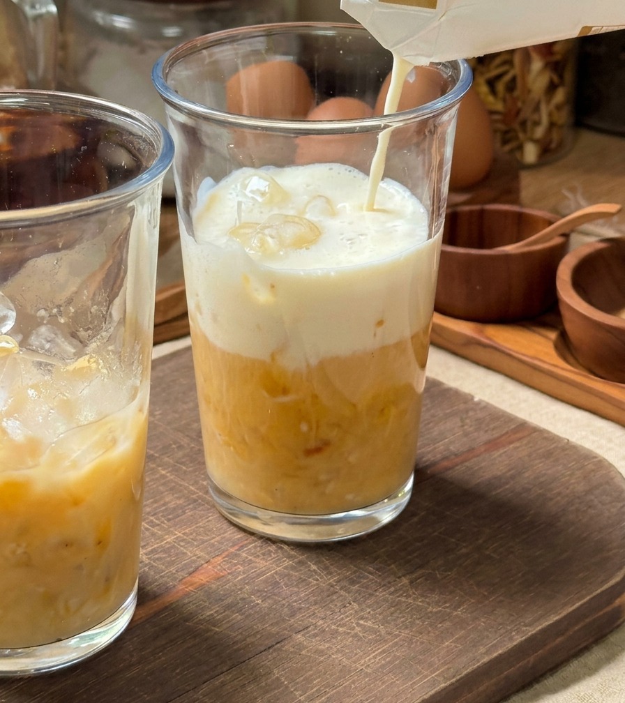 Du lait froid versé depuis une brique dans un grand verre rempli de glaçons et dune base de purée de banane jaune.