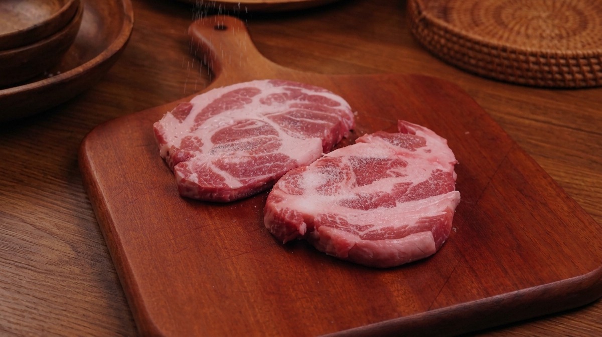 Une main gantée saupoudrant du sel et du poivre sur deux steaks de porc crus posés sur une planche en bois.