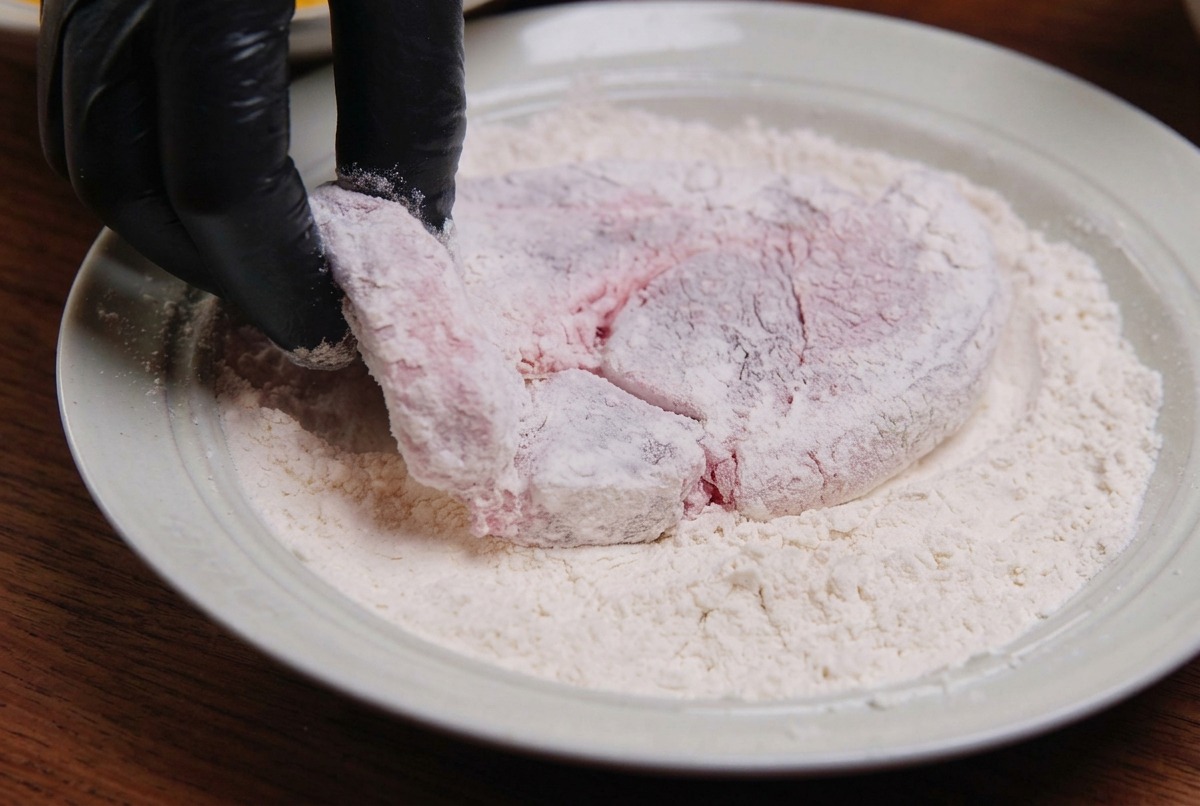 Un morceau de porc cru enrobé de farine blanche dans un plat en céramique blanc par une main gantée.