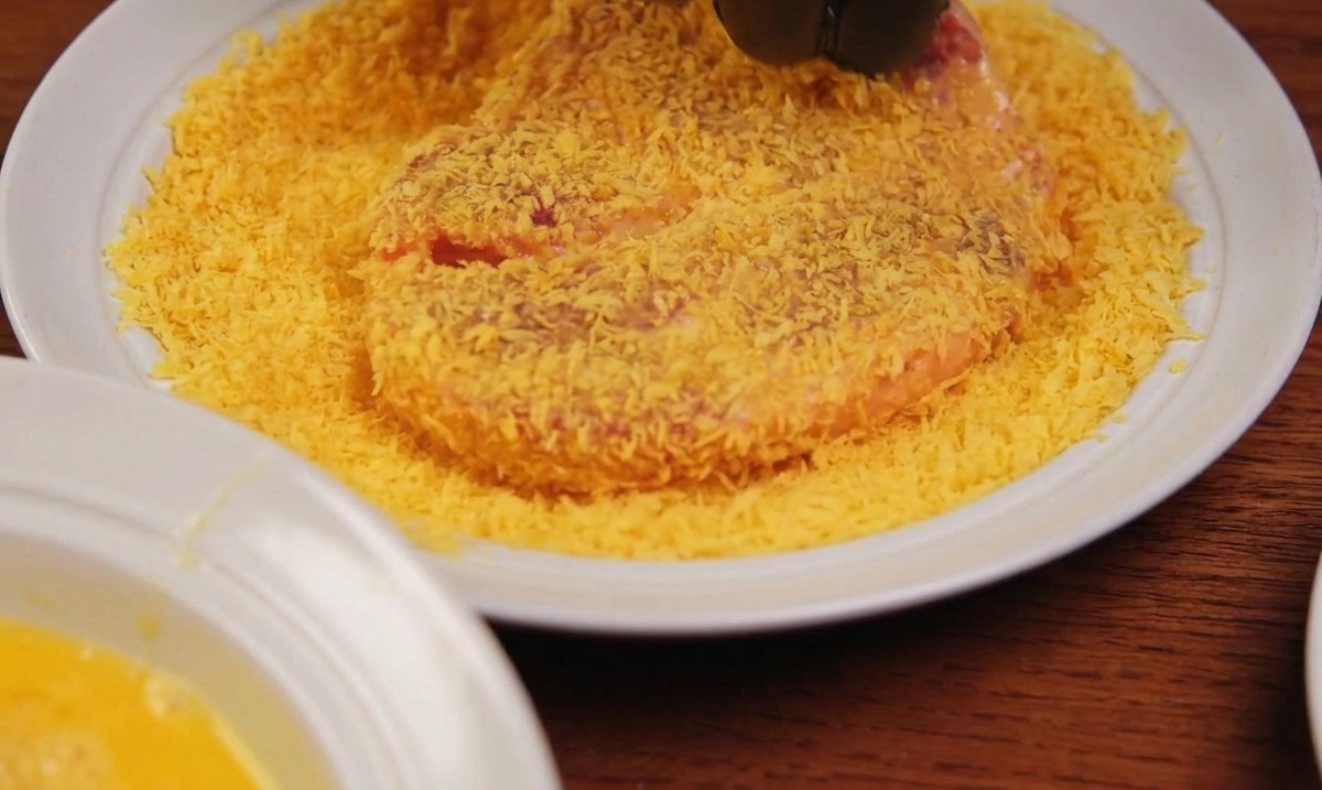 Une main portant un gant noir pressant un morceau de viande dans une assiette recouverte de chapelure panko dorée.