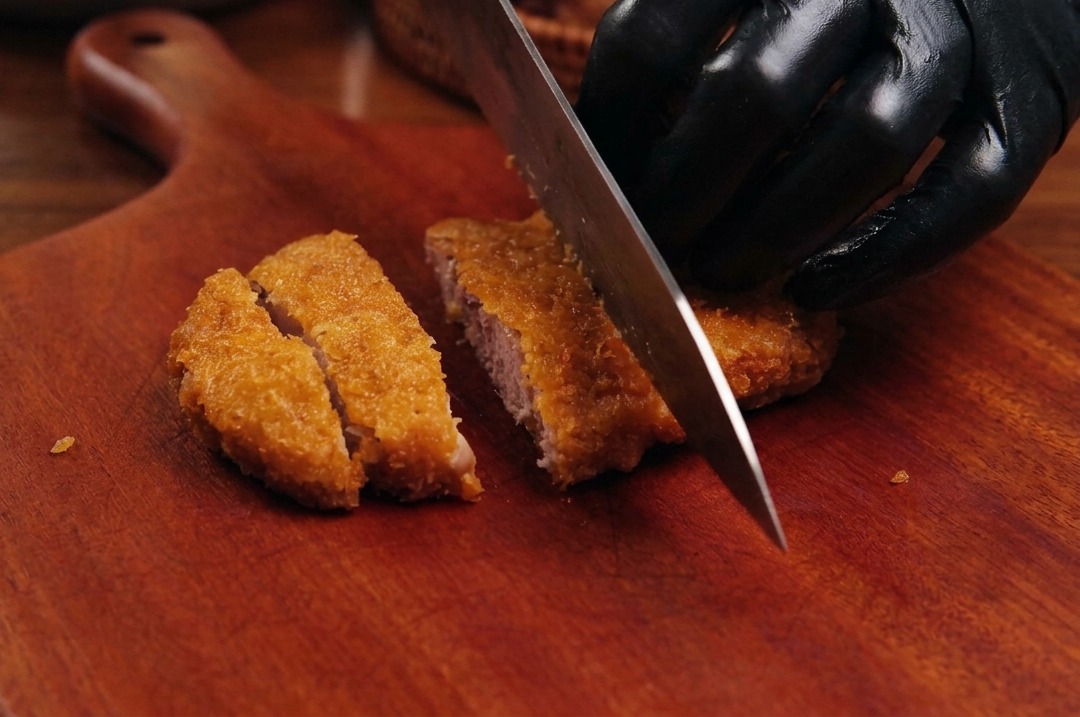 Un couteau de chef tranchant coupant un porc frit et pané bien doré sur une planche en bois.