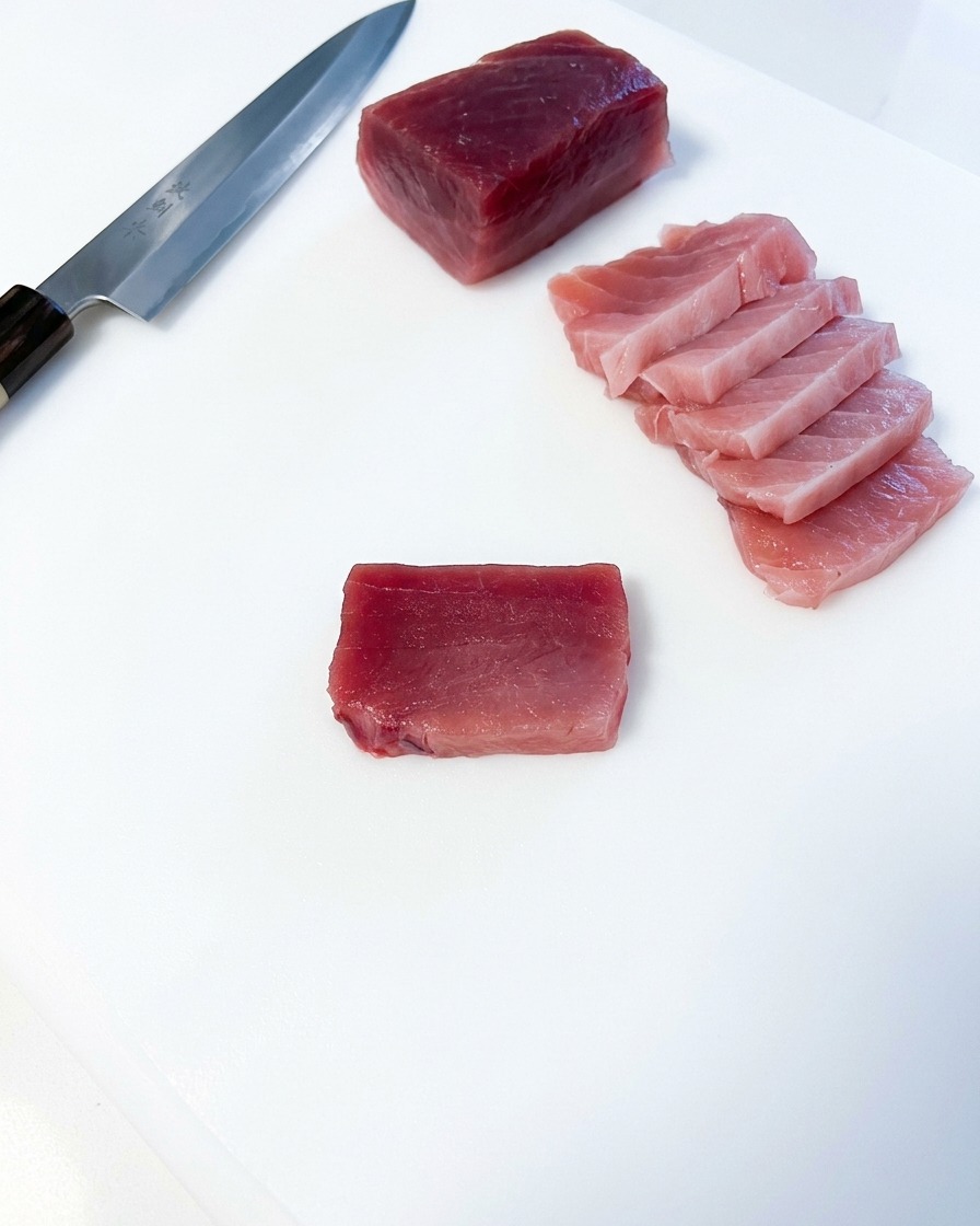 Plusieurs tranches rectangulaires de thon gras rose disposées à côté dun gros bloc de thon et dun couteau de cuisine japonais sur une planche blanche.