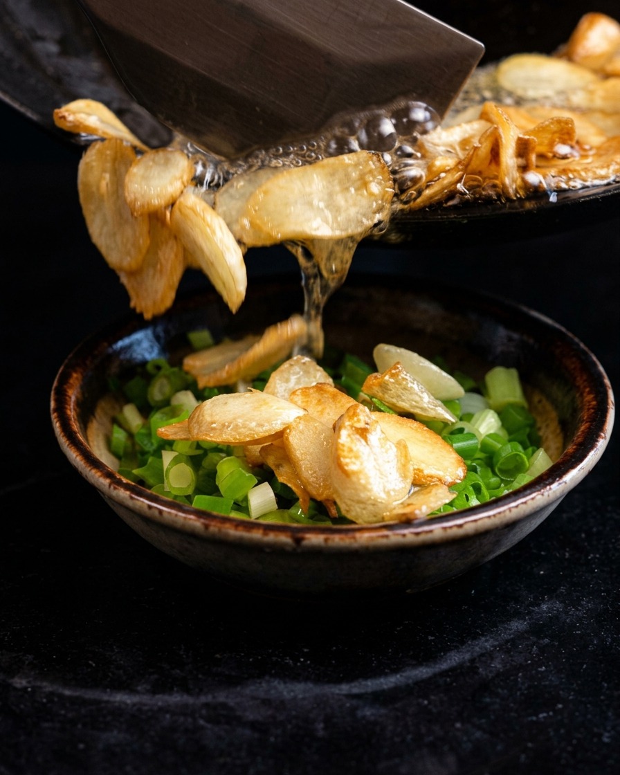 Verser de lhuile bouillante crépitante et des chips dail frit depuis une poêle dans un bol rempli doignons verts émincés.