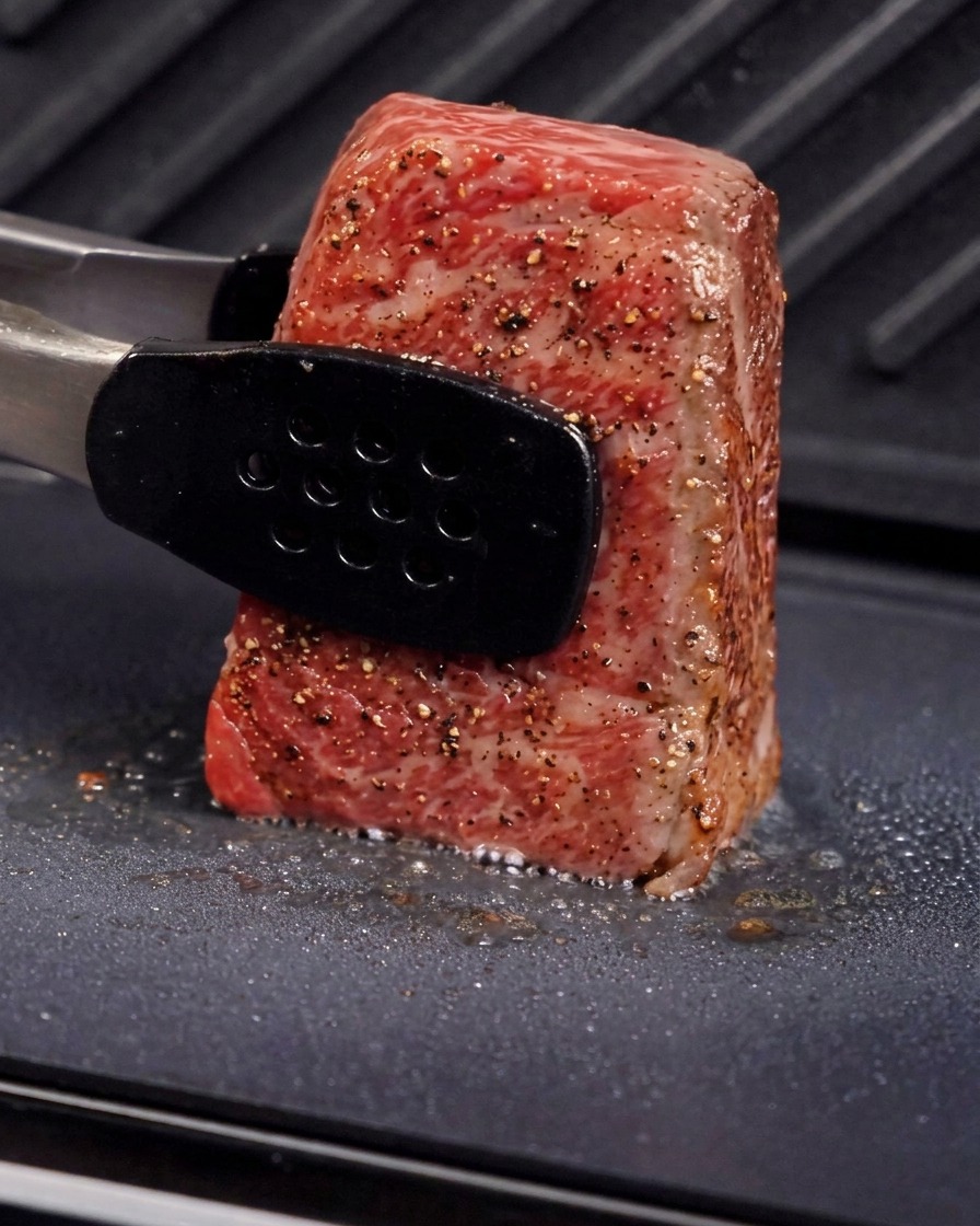 Des pinces métalliques maintenant un morceau épais de bœuf à la verticale sur une plaque de gril pour saisir le côté, avec de la vapeur et de lhuile crépitante visibles.