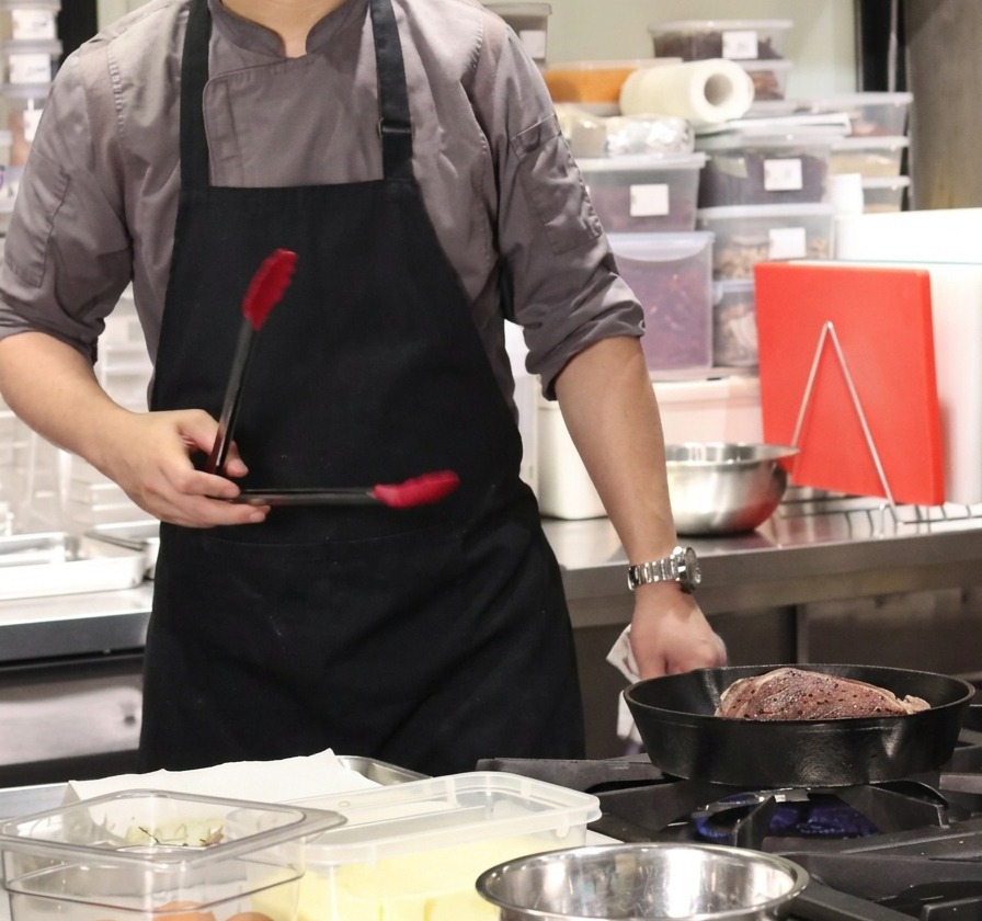 Un chef tenant une pince au-dessus dune poêle en fonte chaude contenant un gros morceau de bœuf pour saisir le gras.