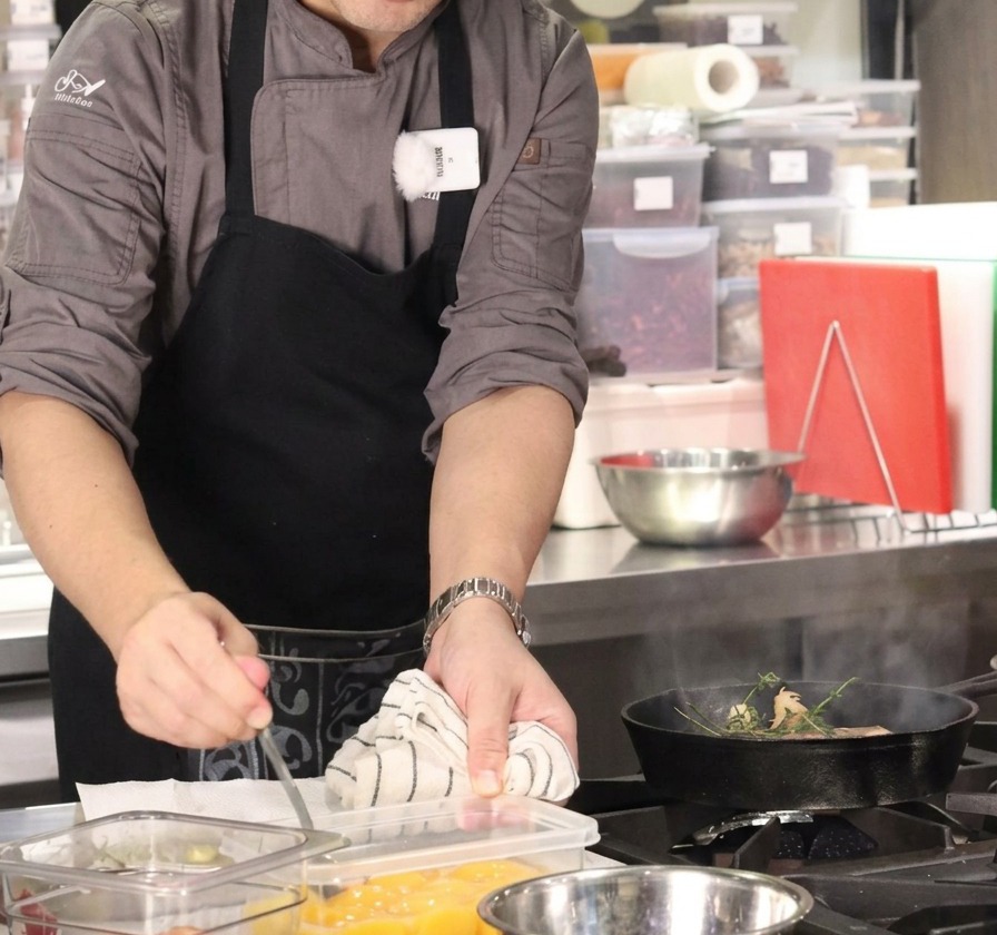 Un chef utilisant une cuillère pour ajouter un cube de beurre dans une poêle fumante avec du bœuf et des herbes.