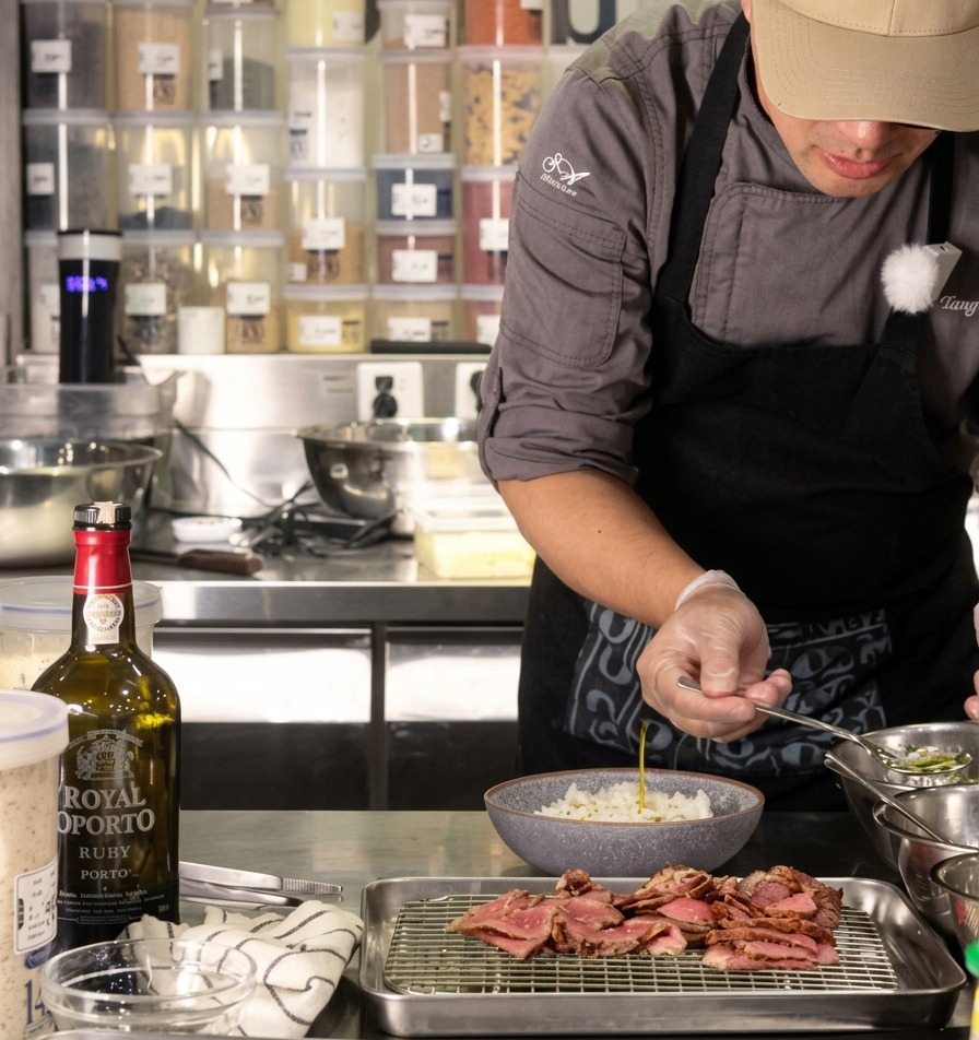 Un chef versant une petite cuillère dhuile doignon vert limpide sur un bol de riz blanc, avec du bœuf tranché reposant sur une grille à côté.