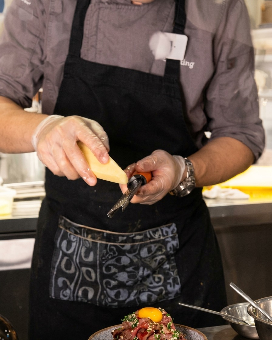 Gros plan dun chef utilisant une râpe microplane pour râper finement du fromage sur un dôme de bœuf tranché surmonté dun jaune dœuf.