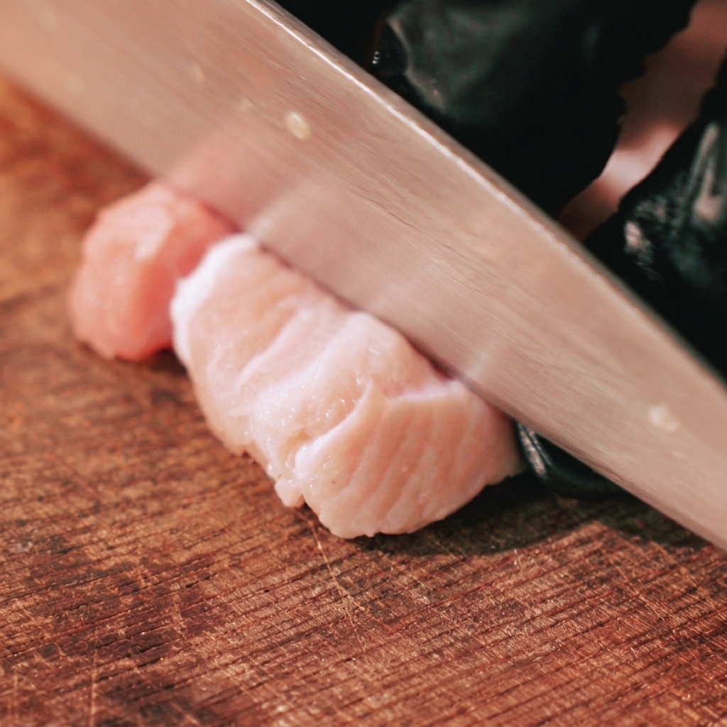 Une main gantée utilisant un couteau de chef pour couper une poitrine de porc crue avec couenne en lanières épaisses sur une planche à découper.