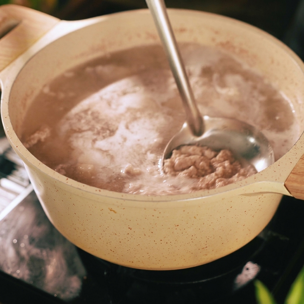 Une louche en métal retirant la mousse grise et les impuretés à la surface de leau bouillante dans une marmite contenant les morceaux de porc.