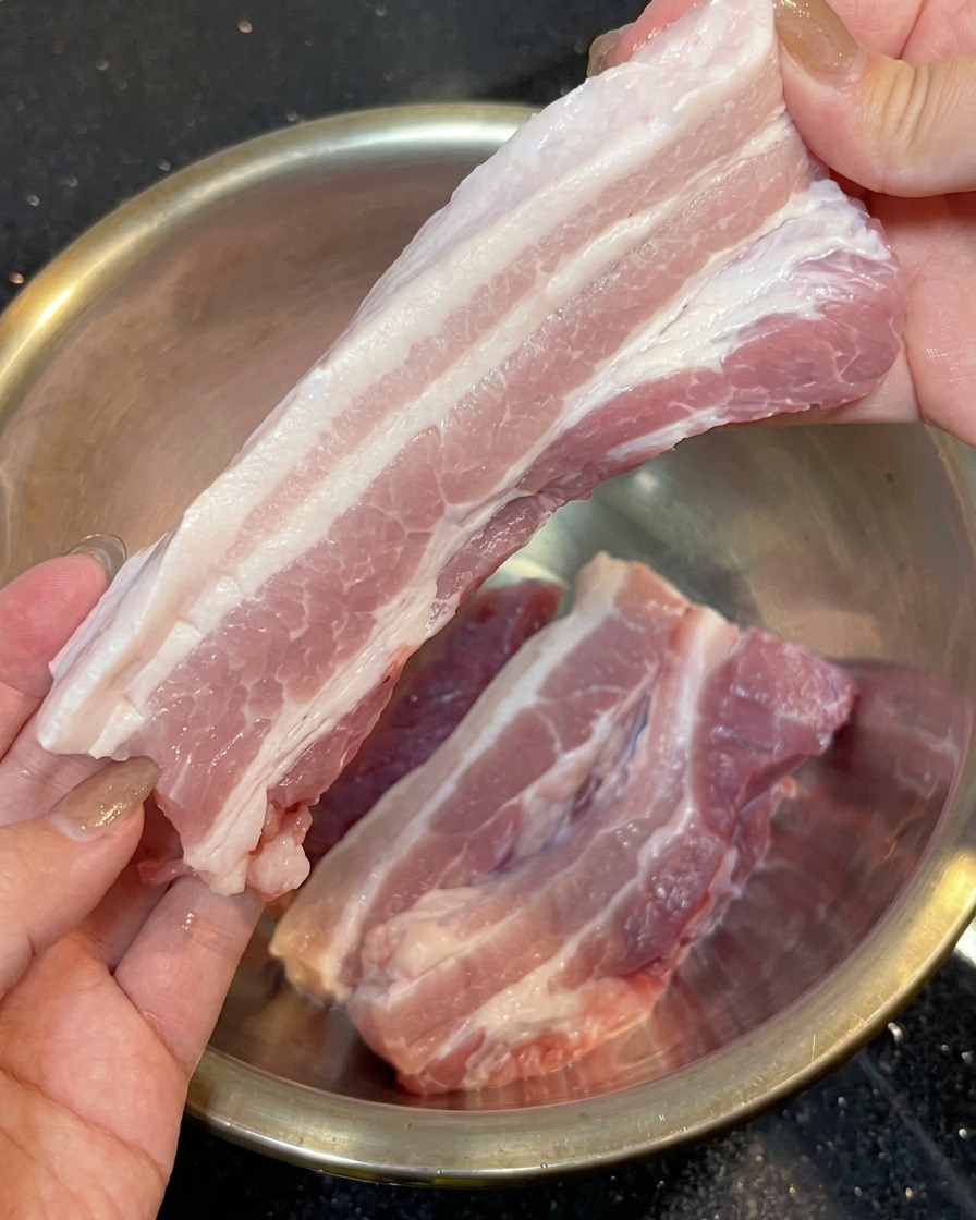 Deux mains tenant une longue tranche de poitrine de porc crue aux couches roses et blanches, sans la couenne, au-dessus dun bol en acier inoxydable.