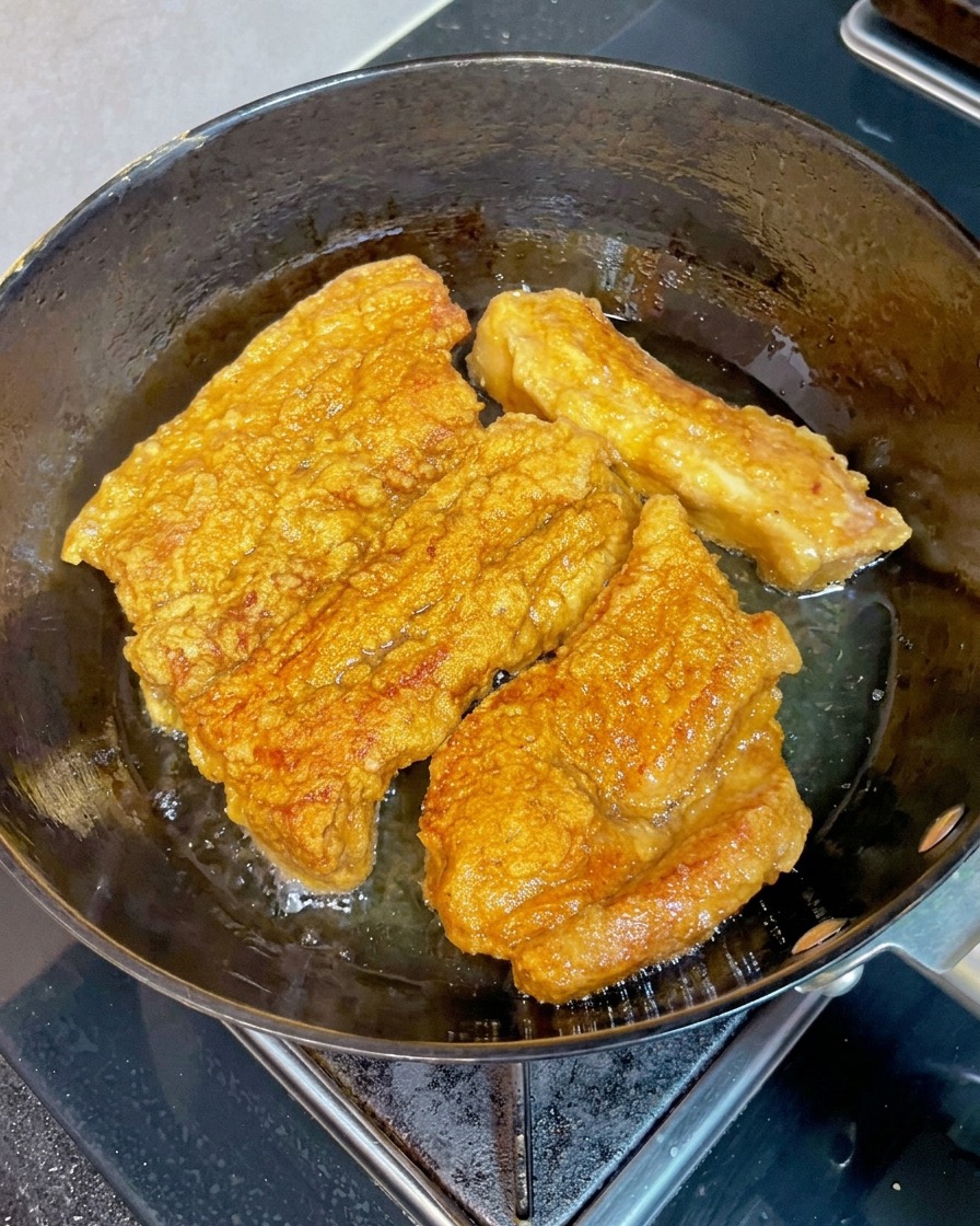 Tranches croustillantes de poitrine de porc brun doré cuisant dans une poêle dhuile chaude, avec une baguette en bois senfonçant dans un morceau pour vérifier sa tendreté.