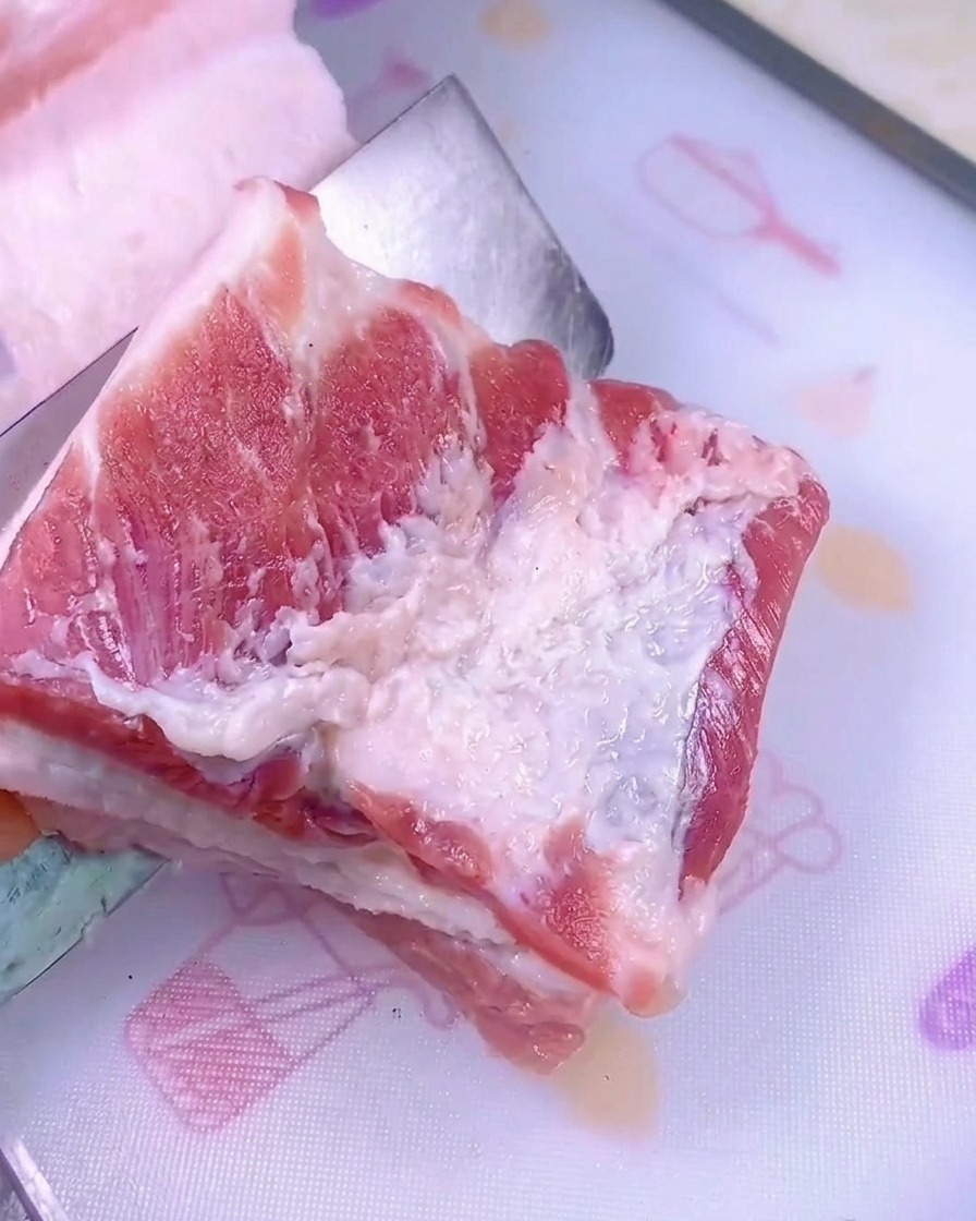 Un couteau bien aiguisé tranchant un morceau de poitrine de porc crue sur une planche à découper en plastique blanc.