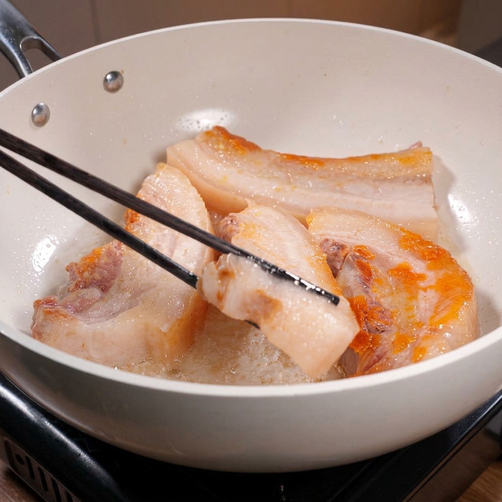 De longues baguettes noires retournant des morceaux de poitrine de porc dans une poêle blanche, révélant des bords bien dorés et croustillants.