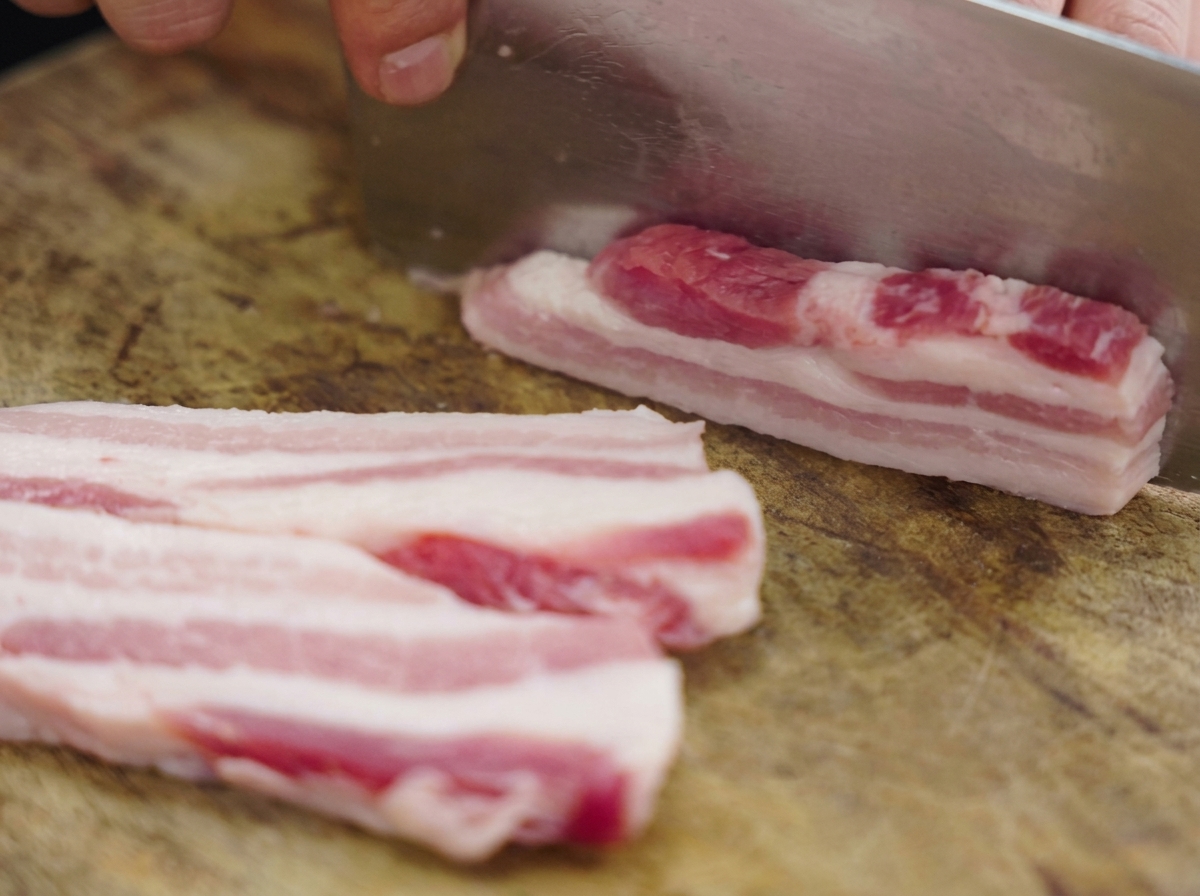 Épaisses lanières de poitrine de porc crue coupées sur une planche en bois avec un grand couperet en métal.