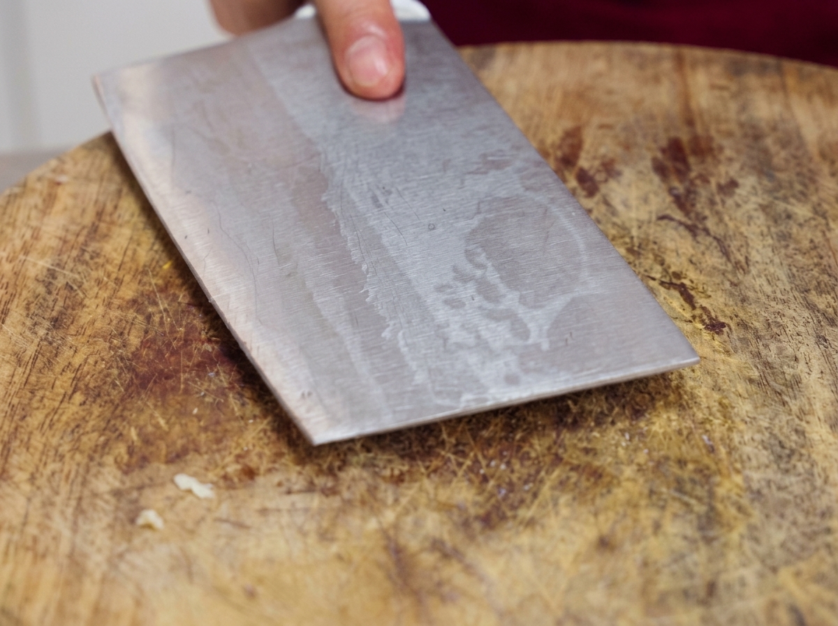 Un couperet en métal lourd pressé à plat sur une planche à découper pour écraser des gousses dail crues.