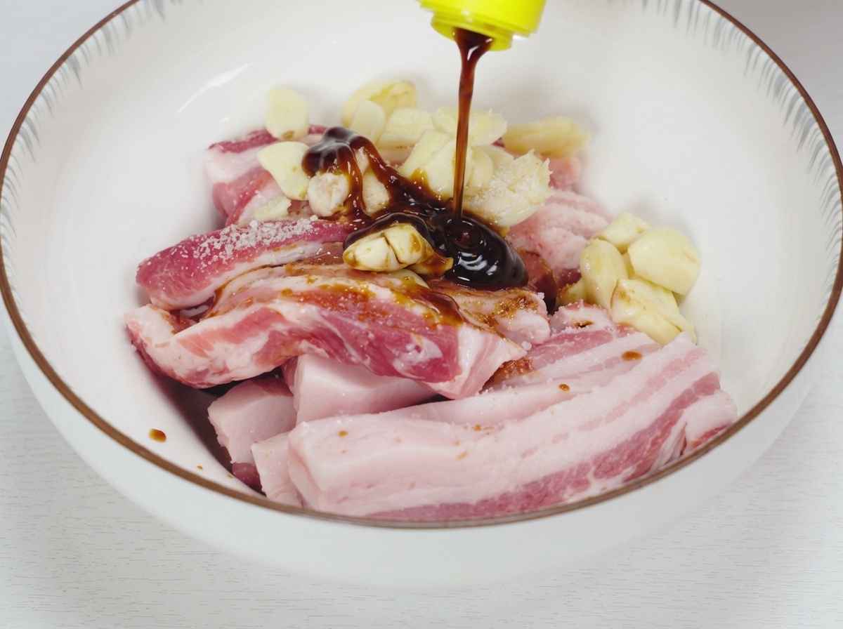 Des sauces liquides sombres et de la sauce dhuître versées sur du porc cru en lanières et de lail écrasé dans un bol en céramique blanc.