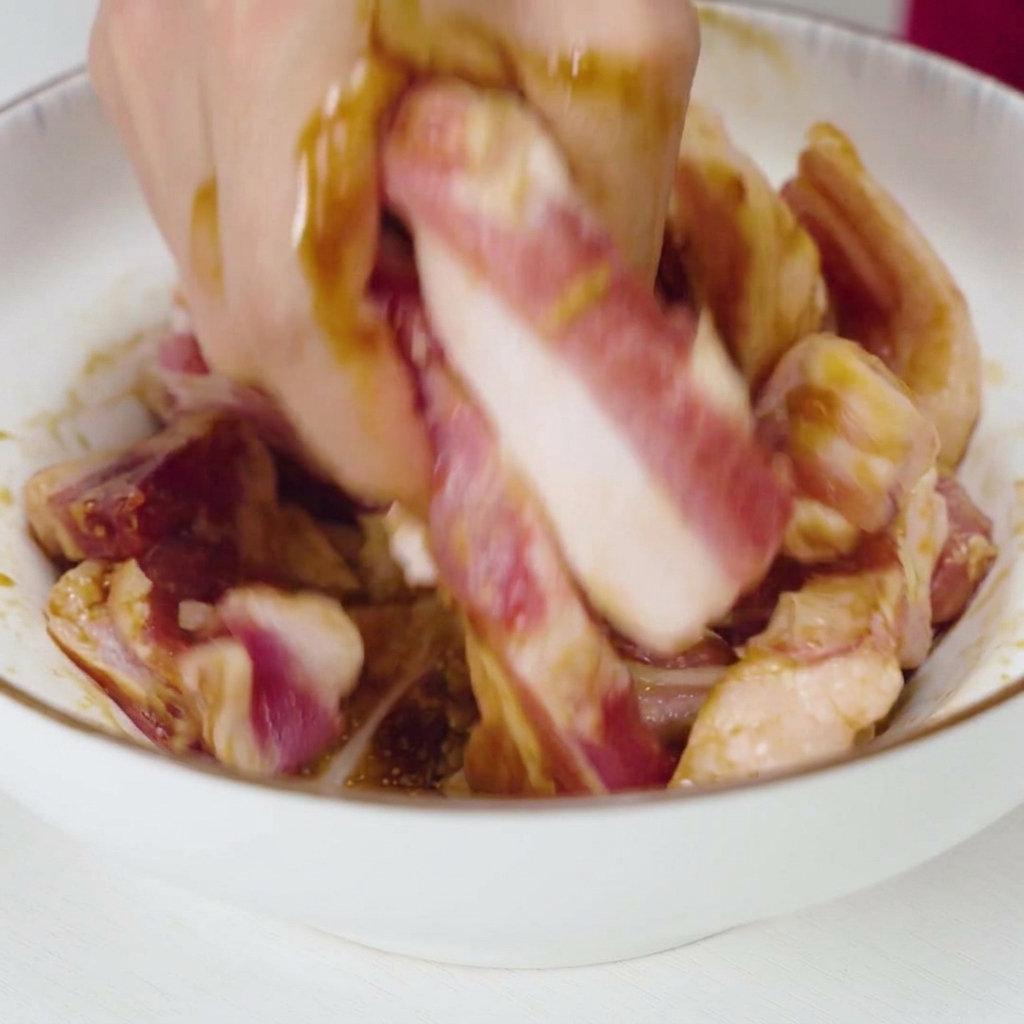 Une main mélangeant et malaxant vigoureusement des lanières de porc cru avec une marinade sombre et de lail dans un bol blanc.