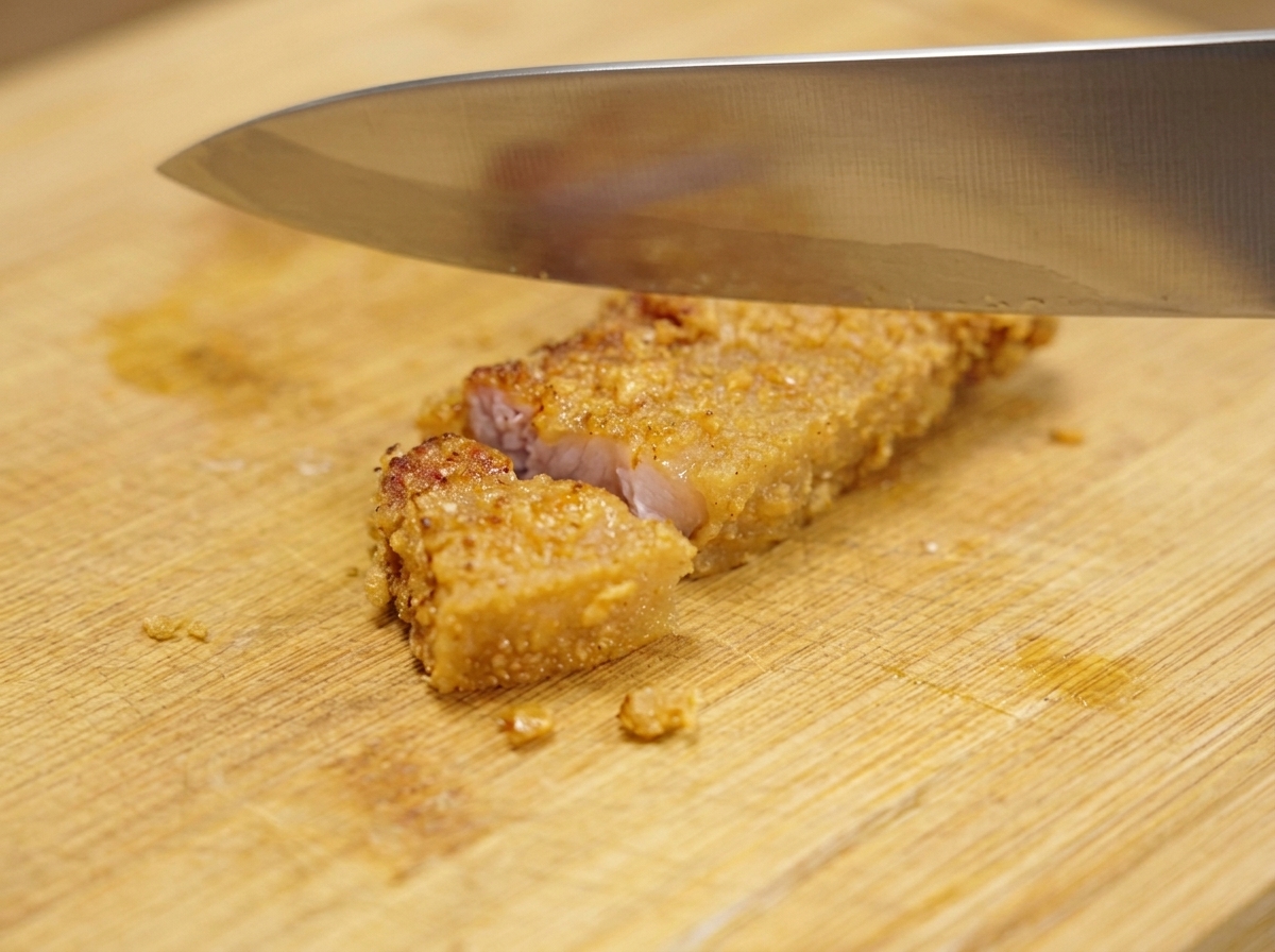 Un grand couteau en métal coupant un morceau de porc frit croustillant en petites sections sur une planche à découper en bois.