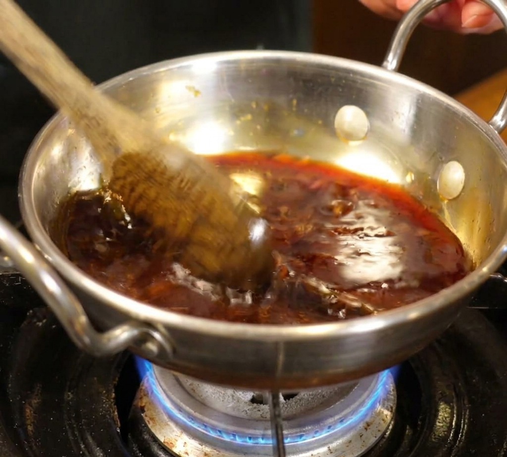 Une cuillère en bois remuant un mélange de sauce foncée qui commence à frémir dans une petite casserole en inox sur une flamme de gaz bleue.