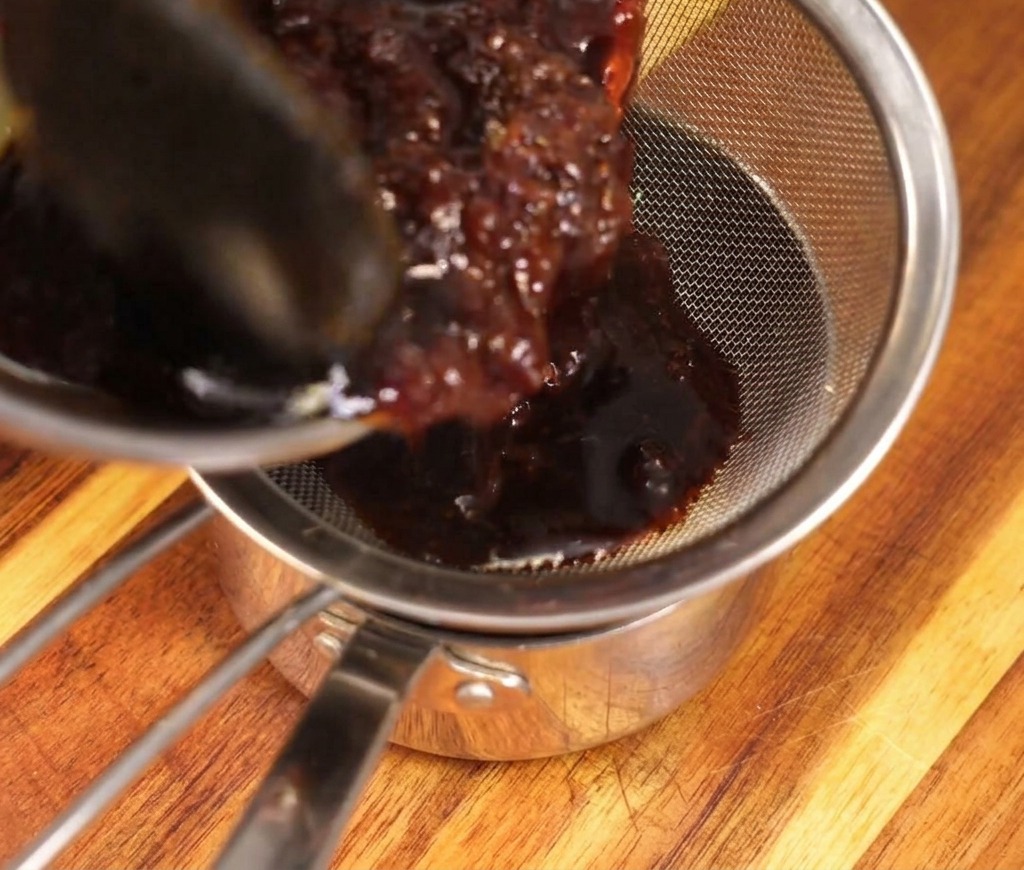 Une sauce riche et foncée versée à travers une passoire fine en métal dans une casserole propre, retenant les flocons de bonite imbibés.