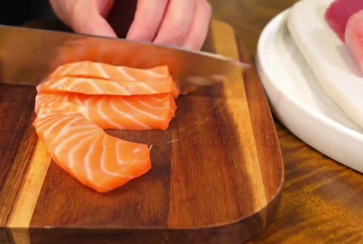 Un couteau de cuisine bien aiguisé coupant un filet de saumon frais et marbré sur une planche en bois, créant des tranches épaisses de sashimi.