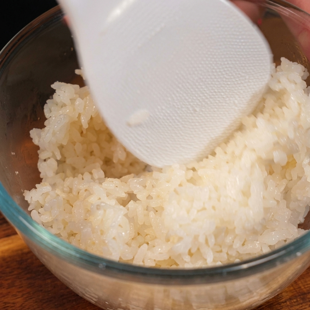 Une spatule à riz en plastique blanc texturé mélangeant délicatement du riz blanc cuit et assaisonné dans un bol en verre transparent.