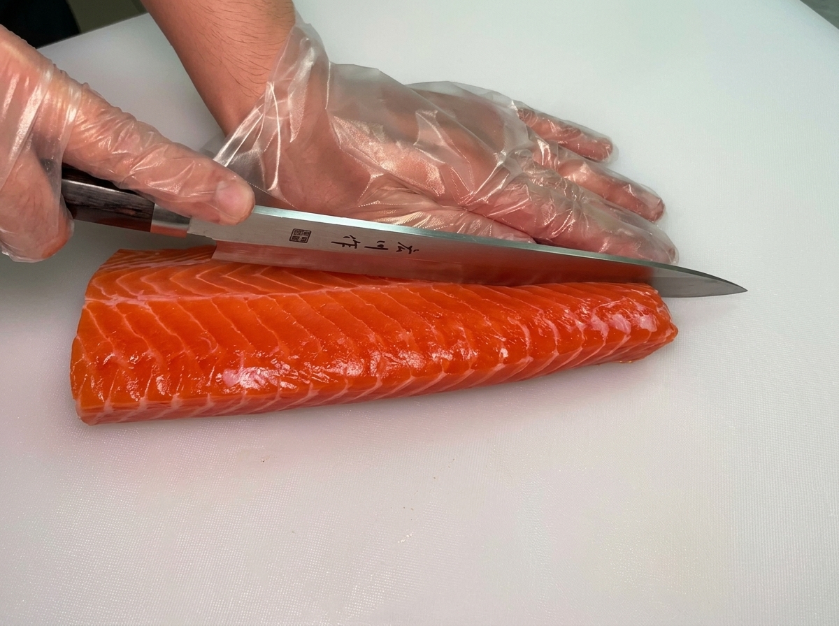 Mains gantées utilisant un long couteau à sashimi pour couper en deux un grand filet de saumon cru orange vif sur une planche à découper blanche.