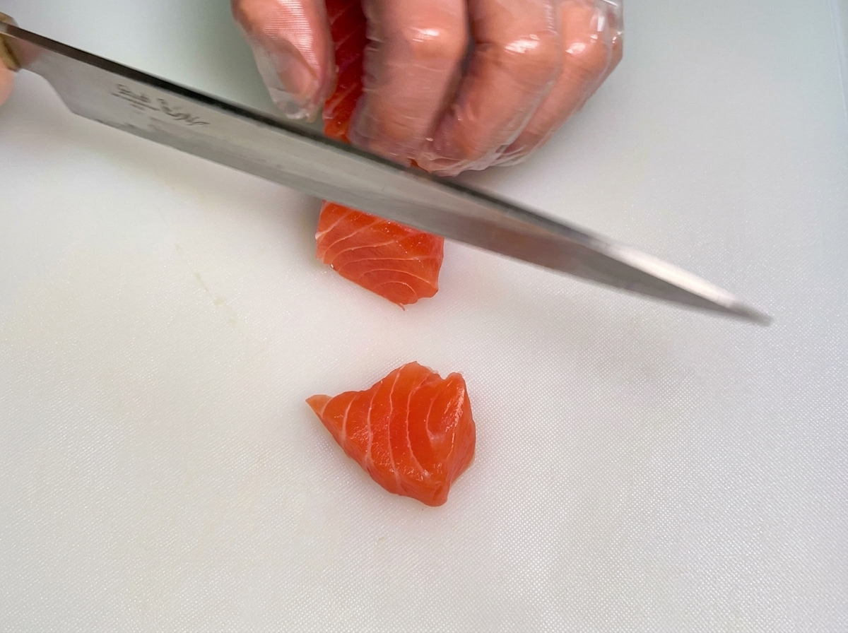 Main gantée dun chef maintenant un morceau de saumon cru pendant que lautre main utilise un couteau pour le couper en bandes épaisses sur une planche blanche.