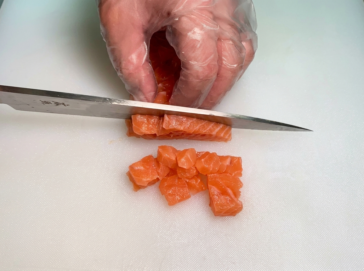 Mains gantées utilisant un couteau pour couper dépaisses bandes de saumon cru orange vif en petits dés uniformes.
