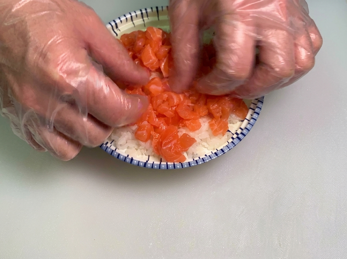 Mains gantées répartissant délicatement une généreuse couche de dés de saumon cru sur un lit de riz à sushi blanc dans un bol aux motifs bleus et blancs.