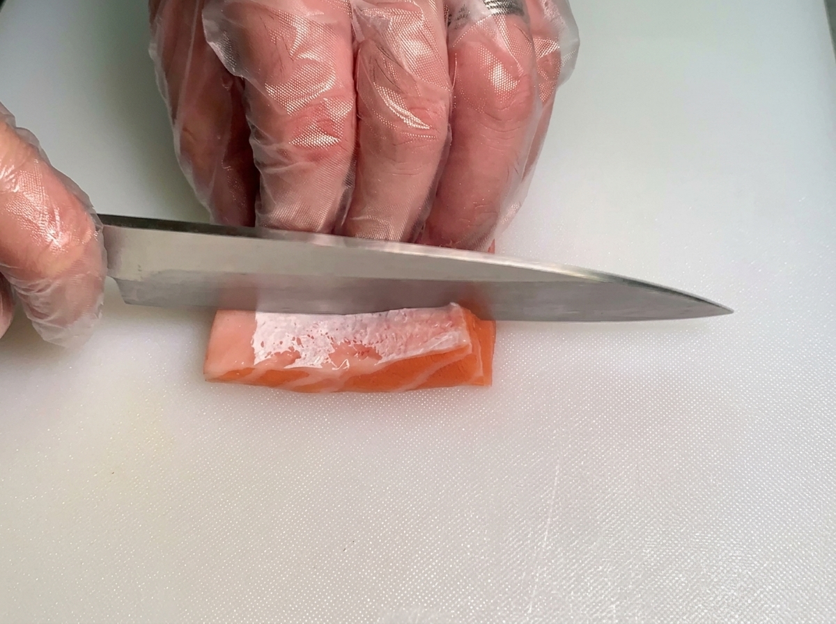 Un chef utilisant un couteau pour trancher délicatement un petit morceau de ventrèche de saumon cru et gras en fines lamelles sur une planche à découper blanche.