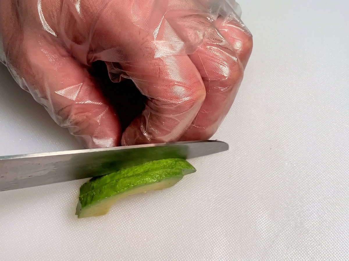 Mains gantées utilisant un couteau pour couper une tranche davocat vert frais en petits cubes.