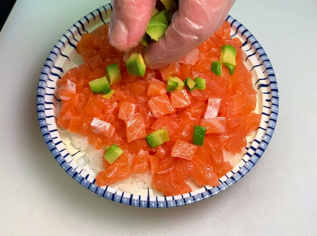 Une main portant un gant transparent parsemant de petits cubes davocat vert sur un bol garni de dés de saumon et de riz blanc.