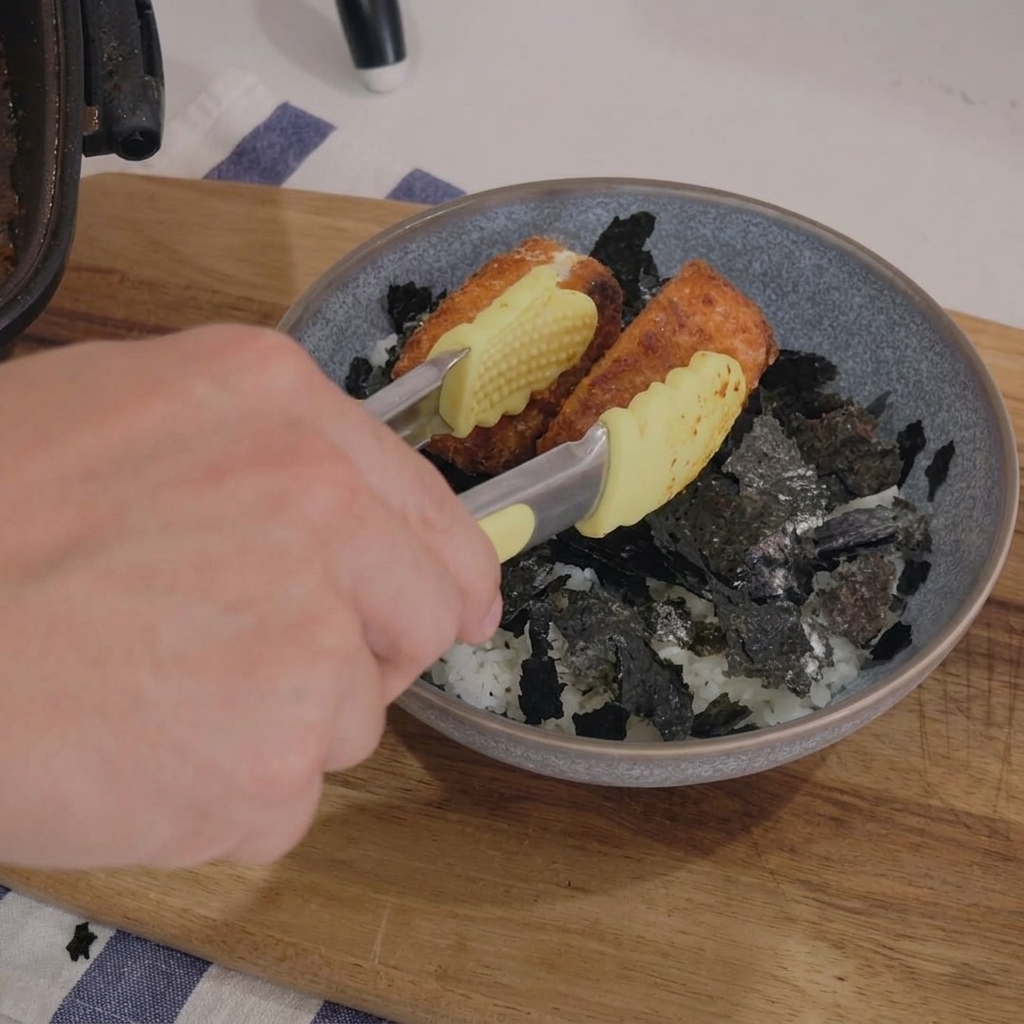 Une main dépose des pavés de saumon dorés à laide dune pince dans un bol en céramique contenant du riz blanc et des morceaux dalgue nori.
