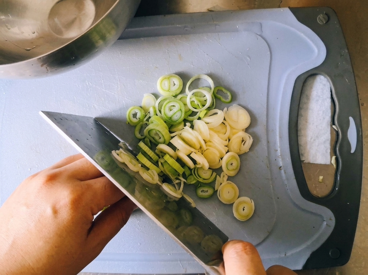 Un chef utilisant un grand couteau de cuisine pour émincer des oignons verts frais en fines rondelles sur une planche bleue.